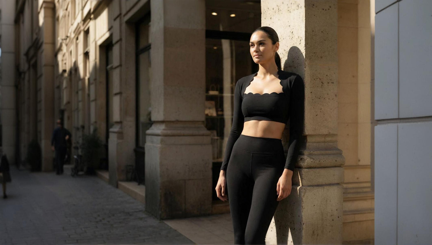 Une photographie de mode lifestyle mettant en scène une mannequin athlétique dans un environnement urbain classique. Elle porte un ensemble de sport noir coordonné, composé d'un top court à manches longues avec une découpe festonnée caractéristique au niveau du décolleté et d'un legging taille haute. La femme est adossée à une colonne massive en pierre de taille, typique d'une architecture européenne. La lumière est latérale et chaleureuse, évoquant une fin de journée, ce qui crée des ombres douces