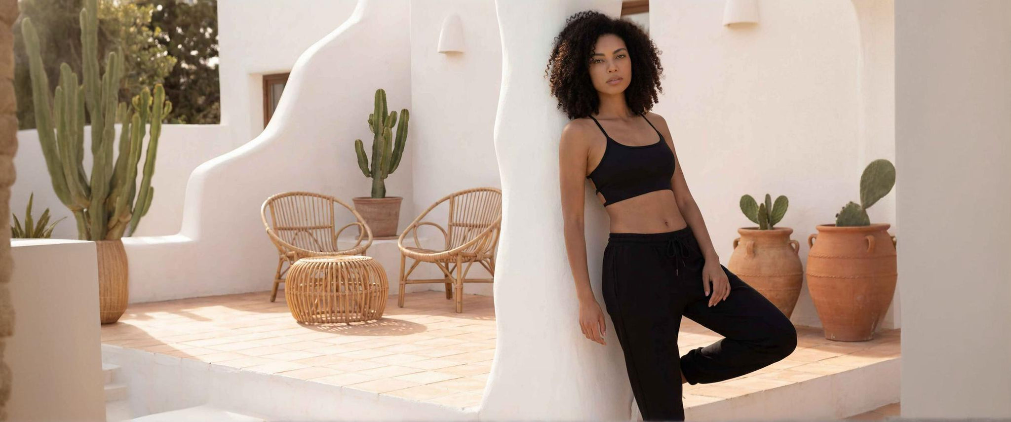 Une femme à la chevelure bouclée pose avec élégance dans un ensemble de sport noir composé de la brassière Mila et d'un bas assorti. Elle s'appuie contre un mur blanc aux courbes méditerranéennes sur un patio décoré de cactus et de mobilier en rotin, créant une atmosphère de bien-être et de sérénité.
