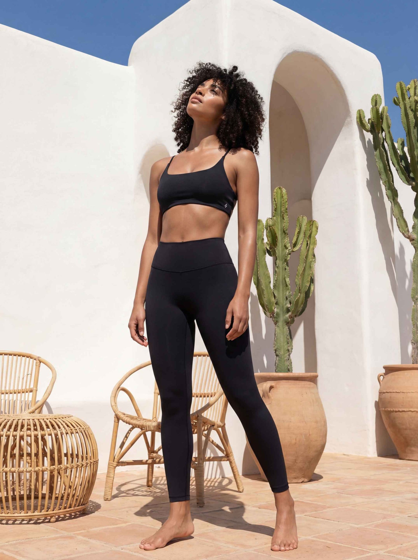 Une mannequin pose en extérieur sous un soleil radieux, vêtue de la brassière Mila noire et d'un legging taille haute coordonné. Le décor présente une architecture méditerranéenne minimaliste aux murs blancs, agrémentée de cactus en pots de terre cuite et de chaises en rotin.