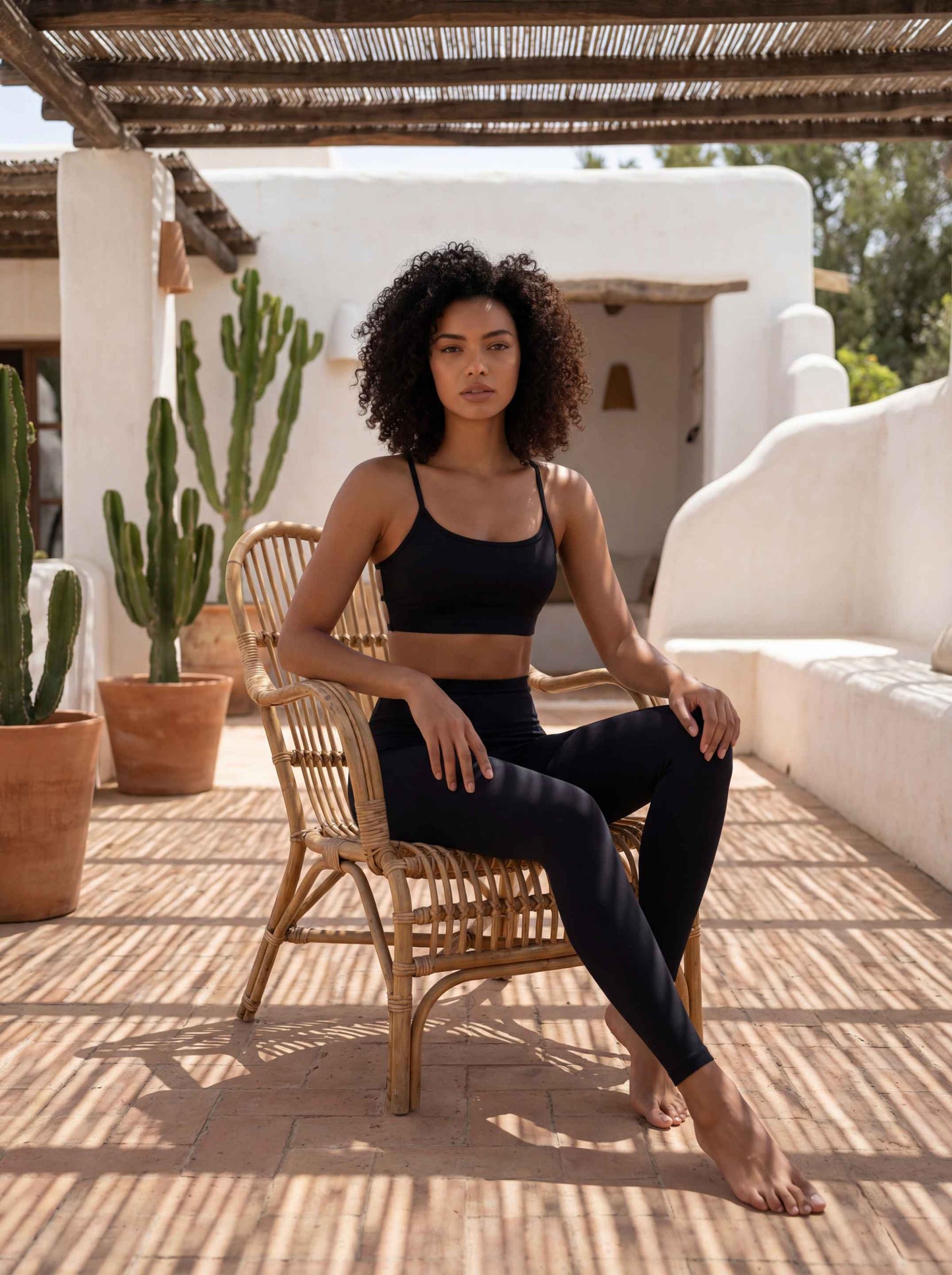 Une mannequin pose dans un ensemble de sport noir élégant composé de la brassière Mila et d'un legging taille haute. Elle est installée dans un décor extérieur minimaliste aux murs blancs, entourée de cactus en pots de terre cuite, sous un jeu d'ombres graphiques créé par une pergola en canisse.