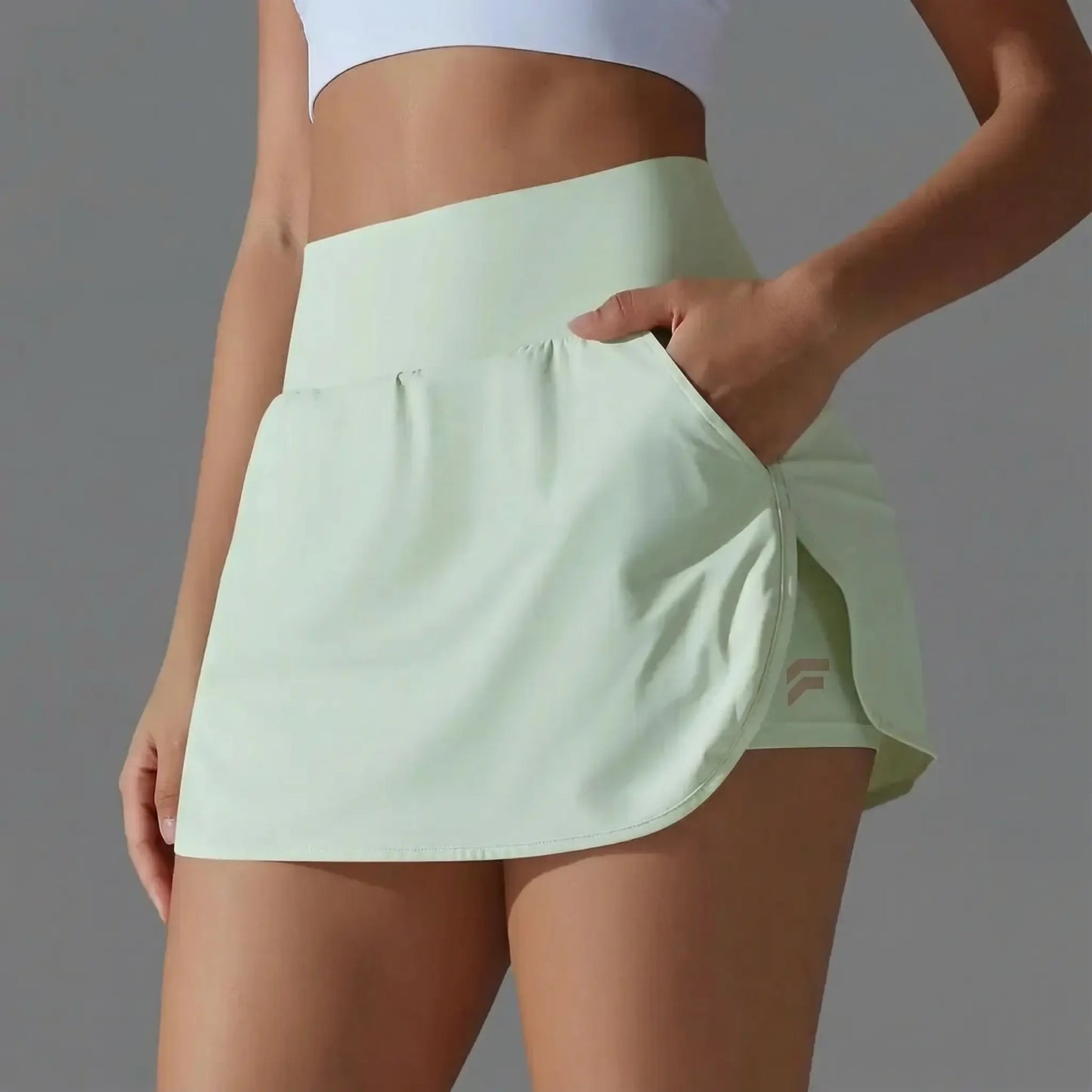 Gros plan d'une femme portant la jupe-short de sport Alma de couleur vert menthe pastel. On voit une main glissée dans une poche latérale fonctionnelle. La jupe présente une taille haute, une coupe fluide avec fente latérale dévoilant le short de compression intégré portant un petit logo discret. Le fond est gris neutre et l'éclairage est de type studio.