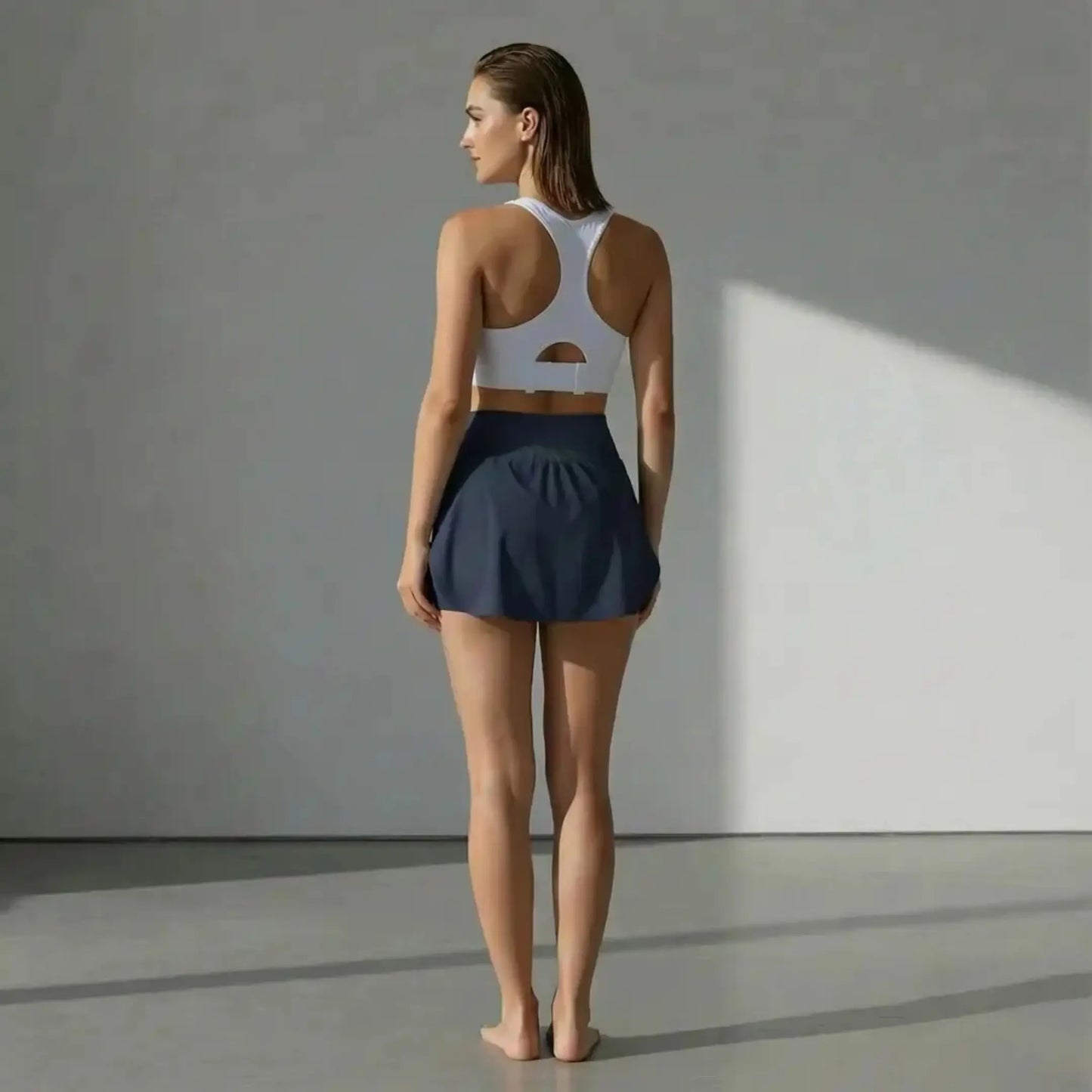 Vue de dos d'une mannequin portant la jupe-short de sport Alma de couleur bleu marine avec une brassière blanche. La coupe est fluide et courte, idéale pour le tennis ou le fitness, présentée dans un studio au design épuré avec un jeu d'ombres naturelles.