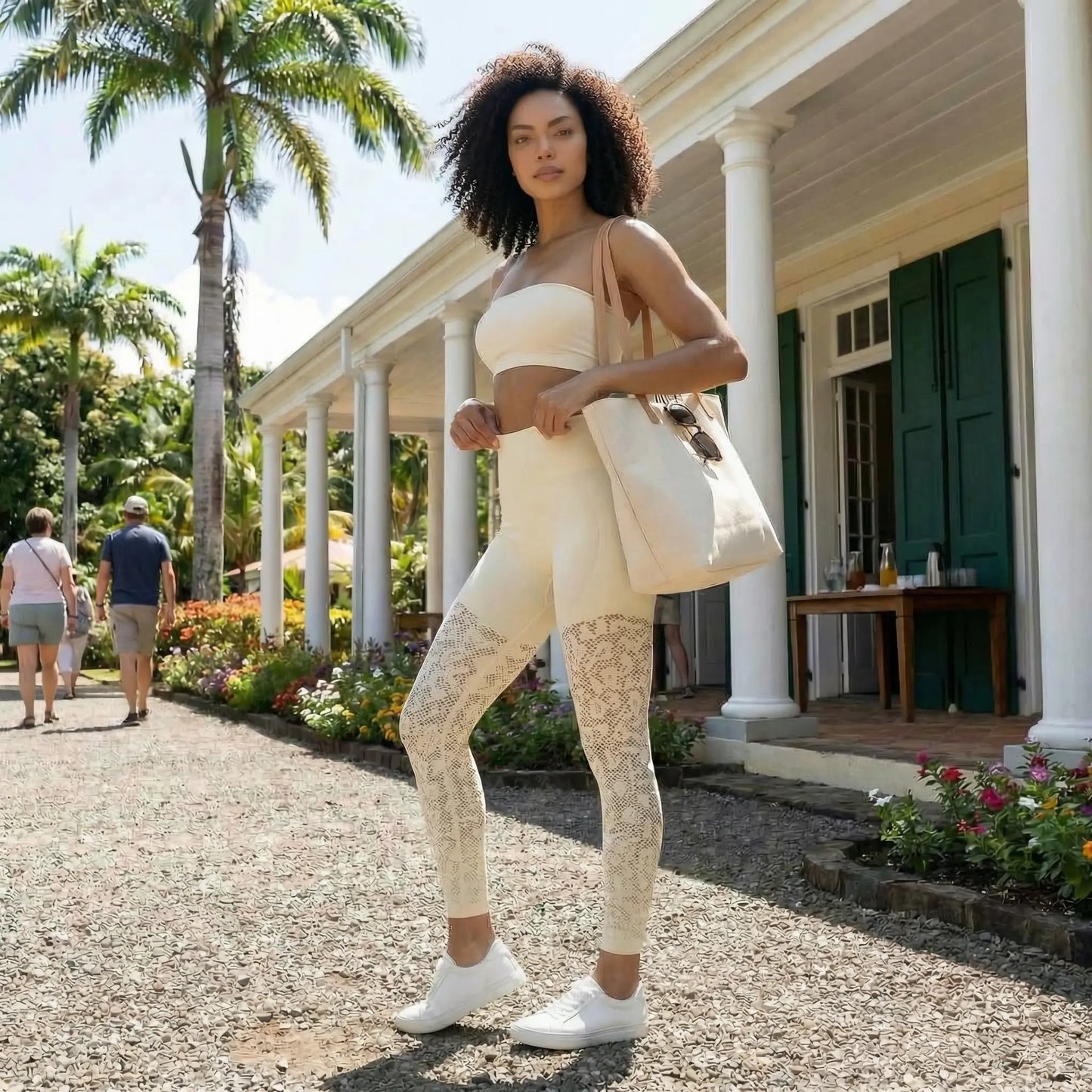 Une mannequin aux cheveux bouclés pose devant une villa coloniale ensoleillée, vêtue du legging galbant Kaia de couleur beige. Le legging se distingue par ses empiècements en dentelle sur les jambes, associé à un haut bandeau, des baskets blanches et un sac cabas en toile.