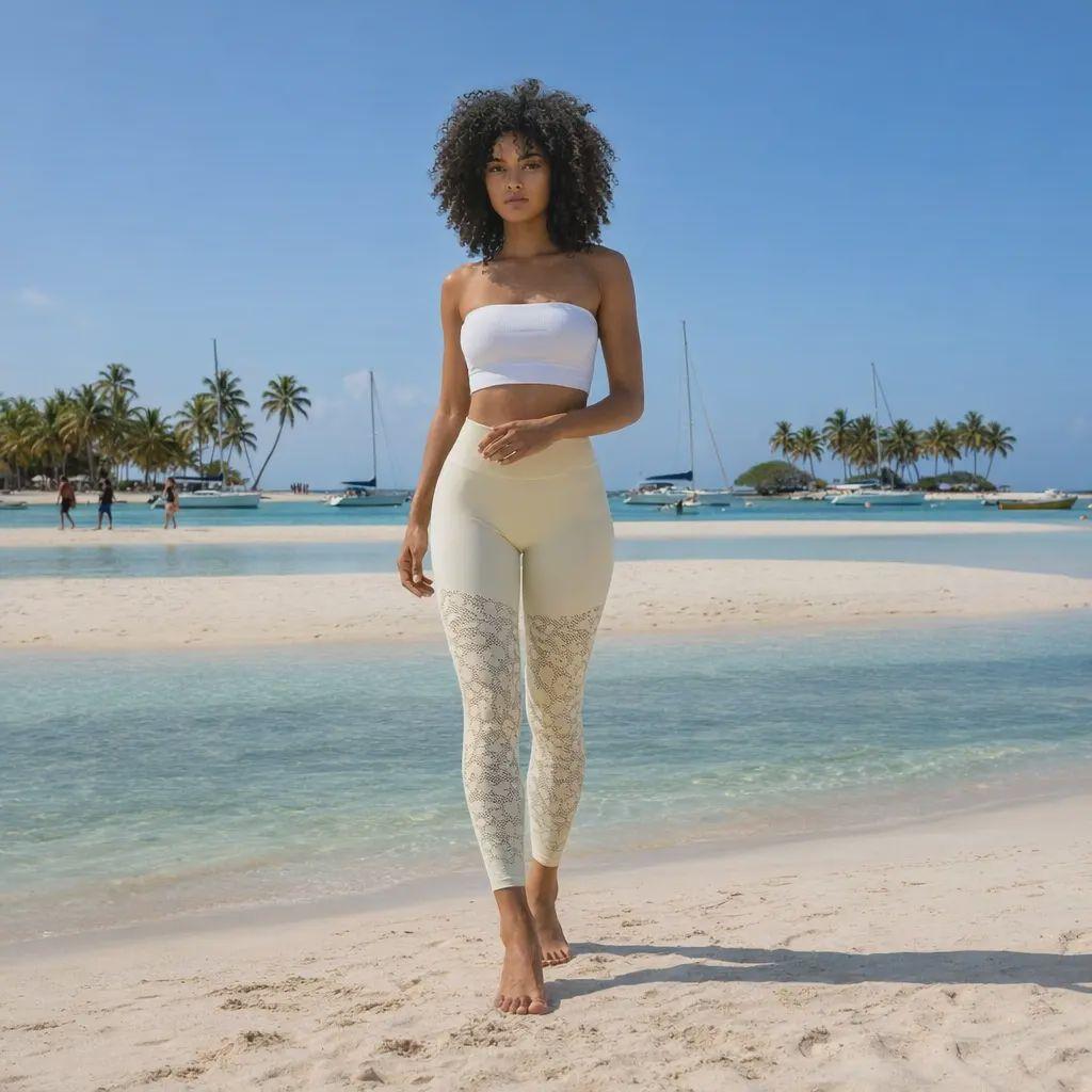 Une jeune femme aux cheveux bouclés pose sur une plage de sable fin, arborant un legging galbant de couleur crème avec des motifs ajourés délicats. Elle complète sa tenue avec un top bandeau blanc, devant un décor paradisiaque composé d'une mer turquoise, de palmiers et de voiliers sous un ciel pur.