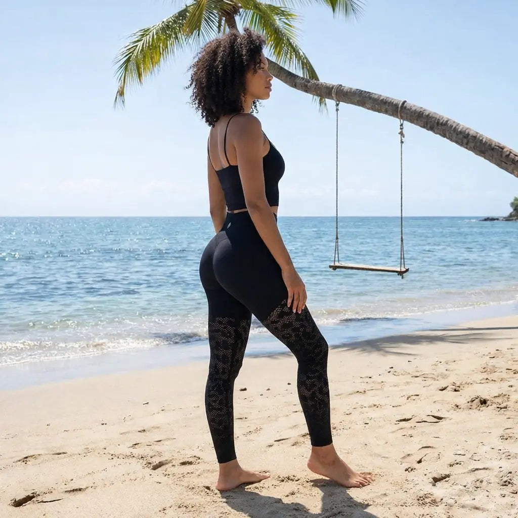Une femme à la chevelure bouclée pose de profil sur une plage de sable fin, mettant en avant un legging galbant noir avec des motifs ajourés sur les jambes. Le décor paradisiaque composé d'une mer turquoise, d'un palmier et d'une balançoire en bois souligne l'aspect lifestyle et bien-être du vêtement.