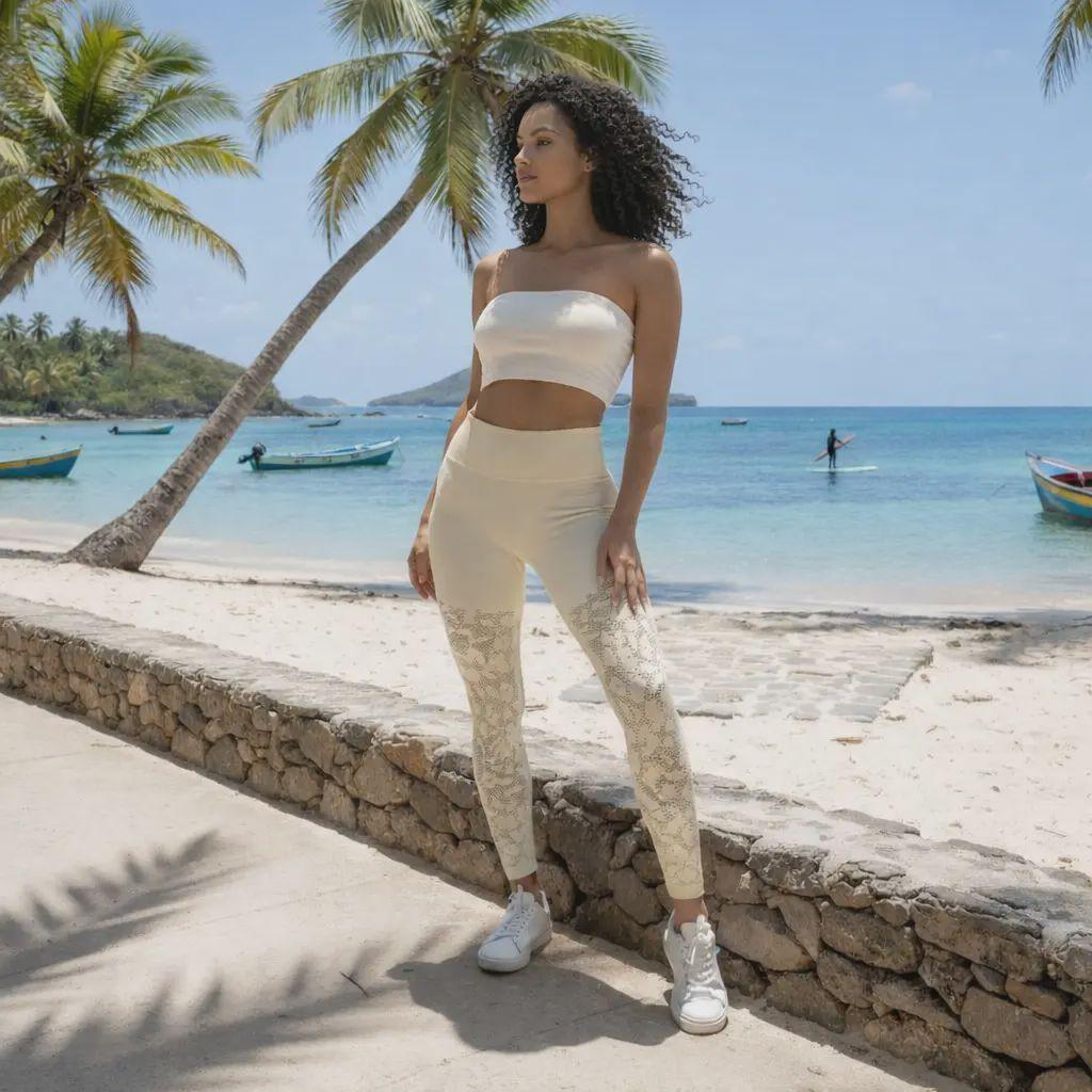 Une mannequin aux cheveux bouclés pose fièrement sur une plage de sable blanc, vêtue d'un legging galbant de couleur crème avec des perforations artistiques aux genoux. Le décor paradisiaque se compose de palmiers inclinés, d'une mer turquoise calme et de petits bateaux de pêche en arrière-plan.