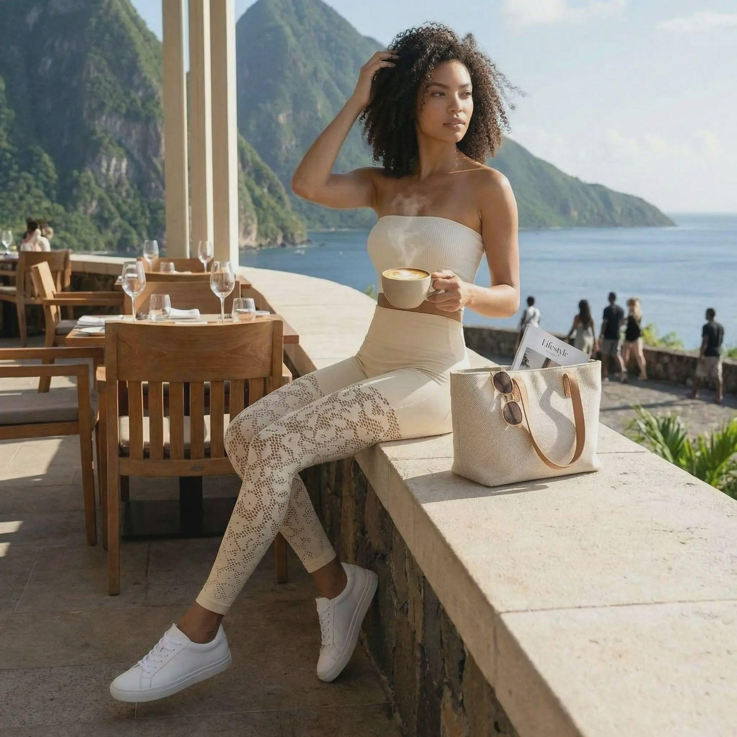 Une femme à la chevelure bouclée pose avec élégance sur un muret en pierre, tenant une tasse de café fumante. Elle porte un legging galbant de couleur crème avec des détails ajourés en dentelle sur les jambes et des baskets blanches, le tout devant un paysage tropical spectaculaire composé de montagnes et de l'océan.