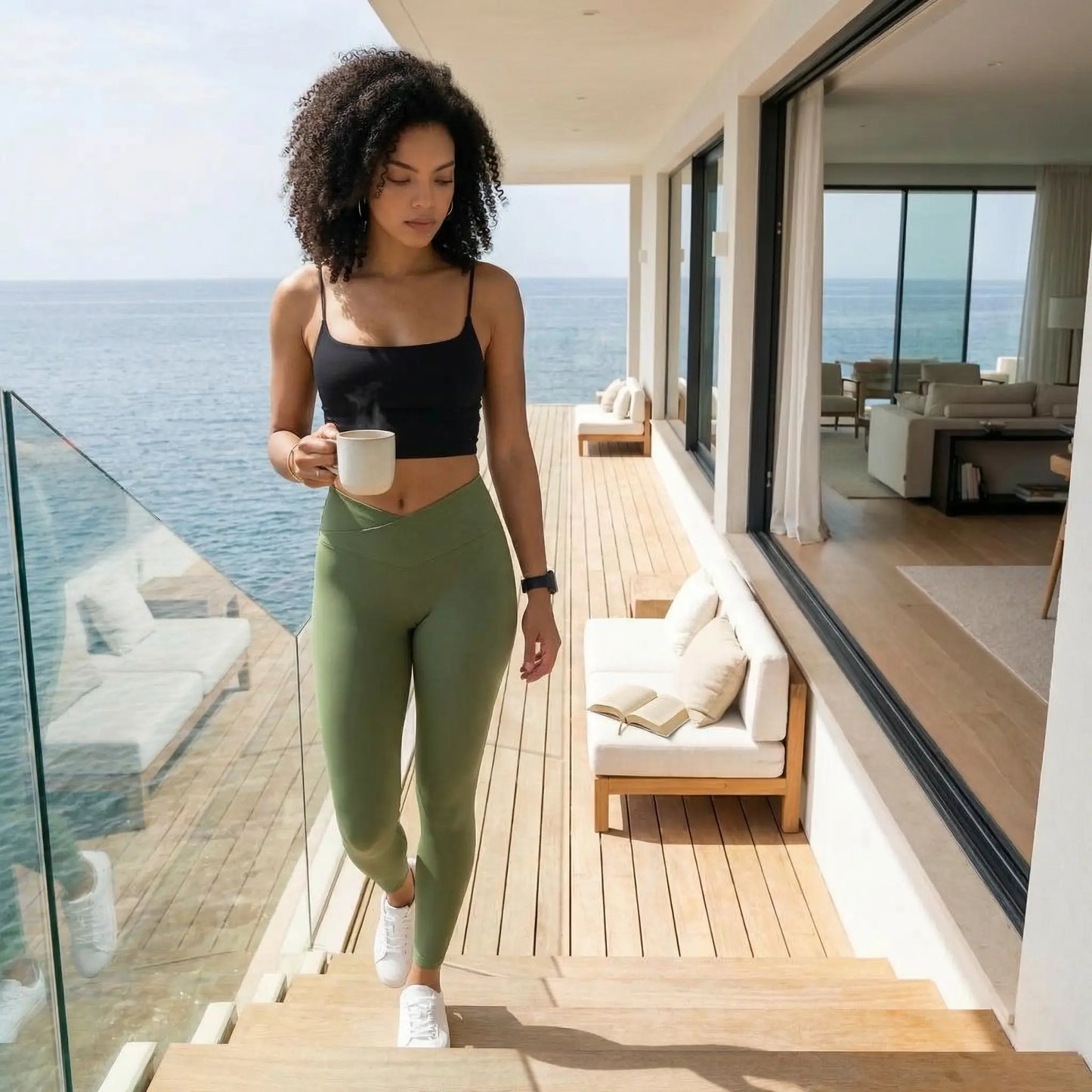 Une jeune femme aux cheveux bouclés porte le legging Elia vert olive à taille croisée et un crop top noir. Elle marche sur le pont en bois d'une villa moderne face à l'océan, tenant une tasse fumante dans un cadre lumineux et apaisant.