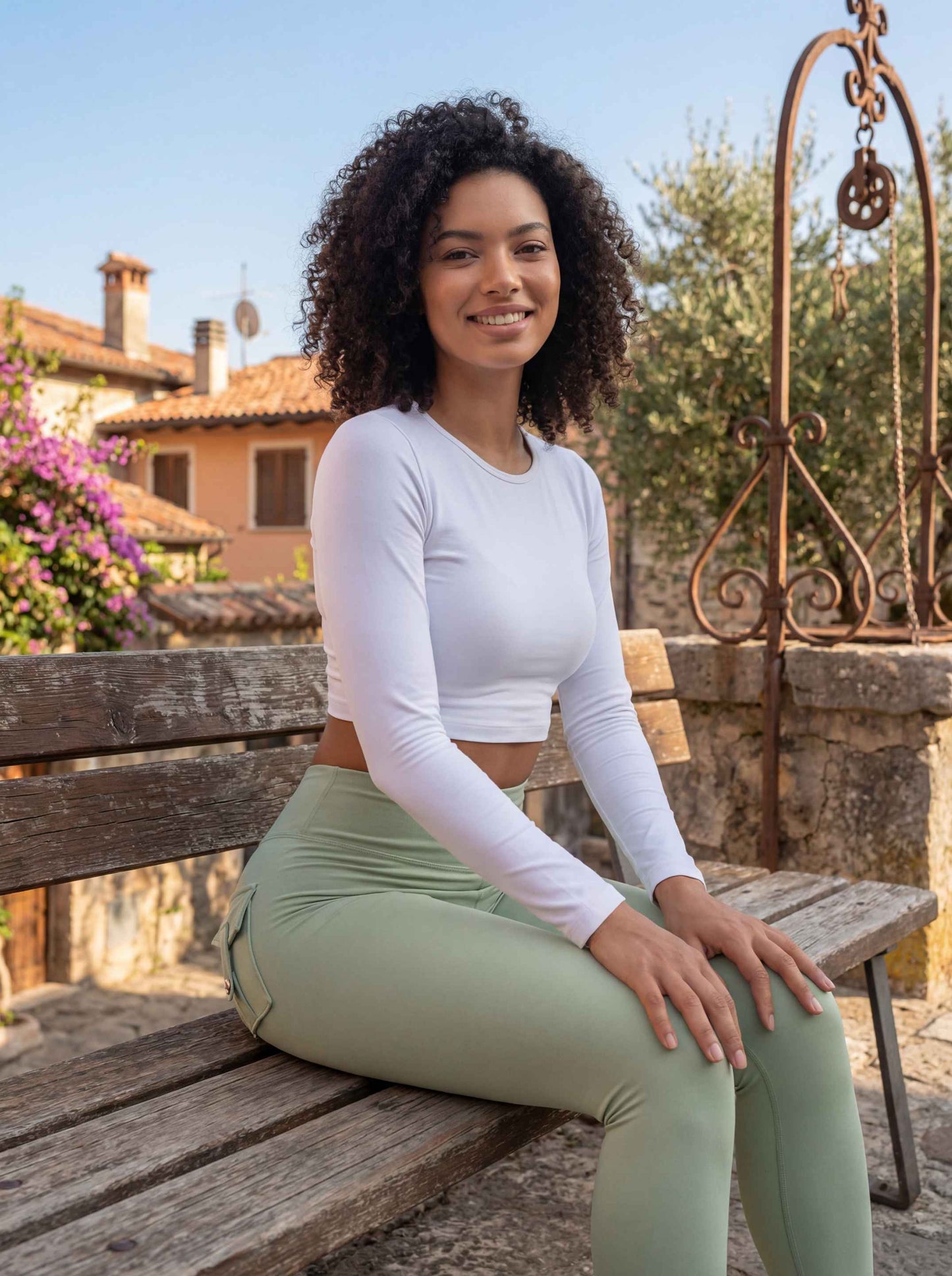Une jeune femme aux cheveux bouclés pose avec assurance, assise sur un banc rustique dans un village pittoresque. Elle porte un legging push up femme de couleur vert sauge mettant en valeur sa silhouette, associé à un top blanc ajusté à manches longues.