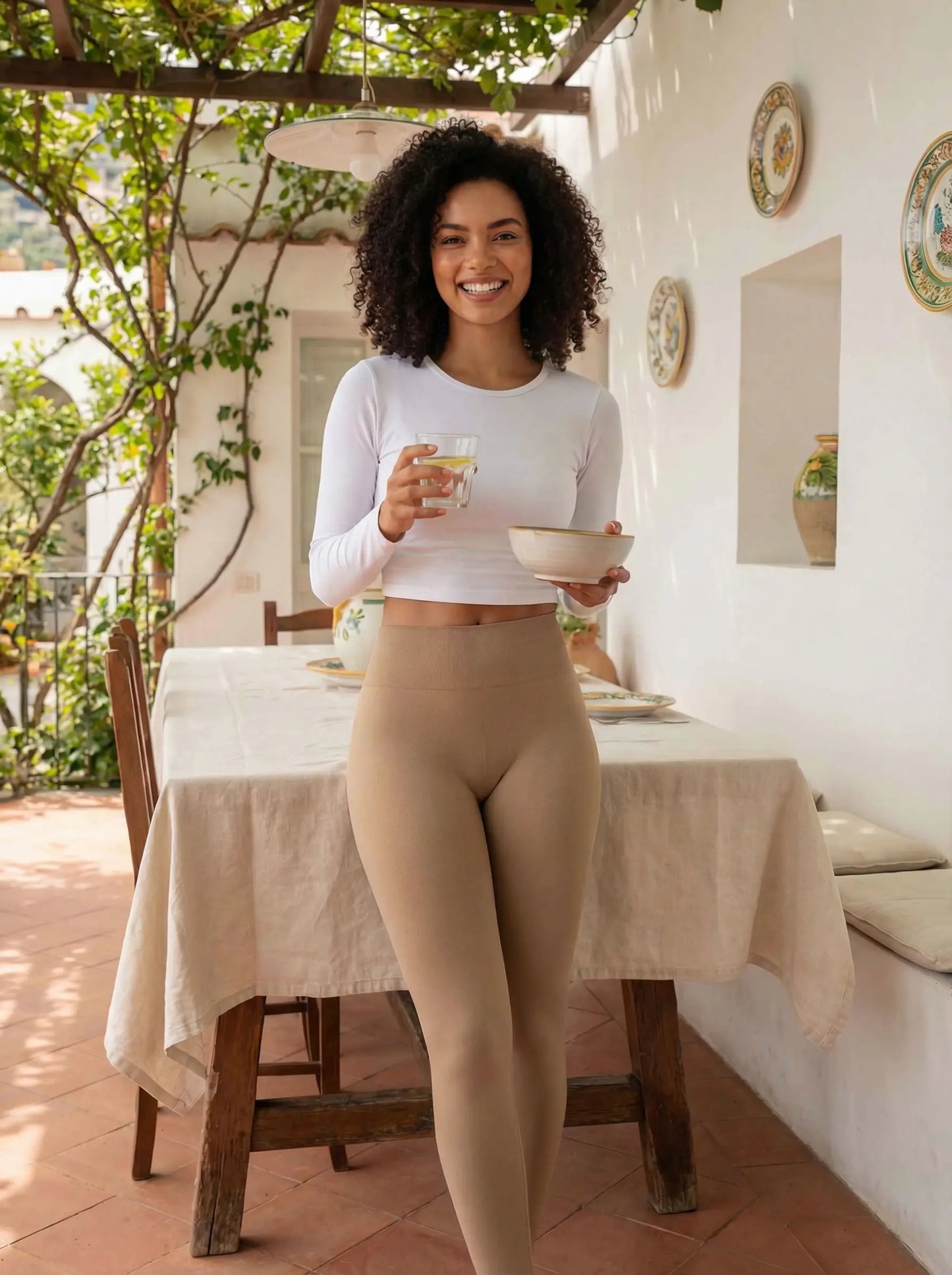 Une jeune femme à la chevelure bouclée pose avec assurance dans un legging push up femme beige galbant. Elle se tient debout près d'une table sur une terrasse ensoleillée, illustrant un mode de vie sain et élégant avec un bol et un verre d'eau à la main.