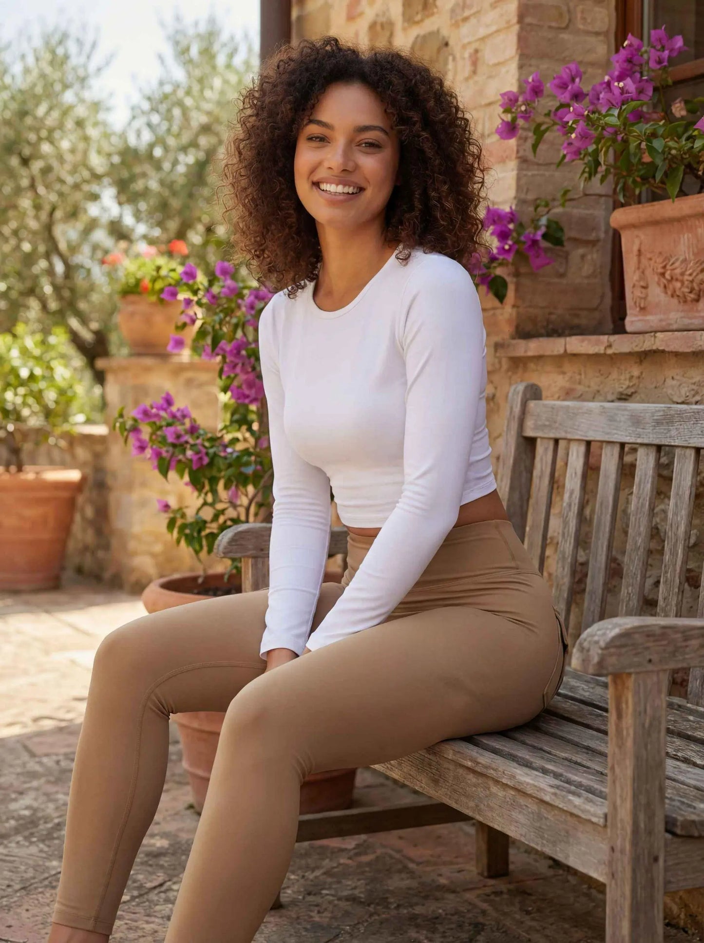Une jeune femme à la chevelure bouclée pose en extérieur sur un banc en bois rustique. Elle porte un legging push up femme de couleur beige/camel qui sculpte la silhouette, complété par un haut blanc ajusté à manches longues dans un décor de jardin méditerranéen ensoleillé.