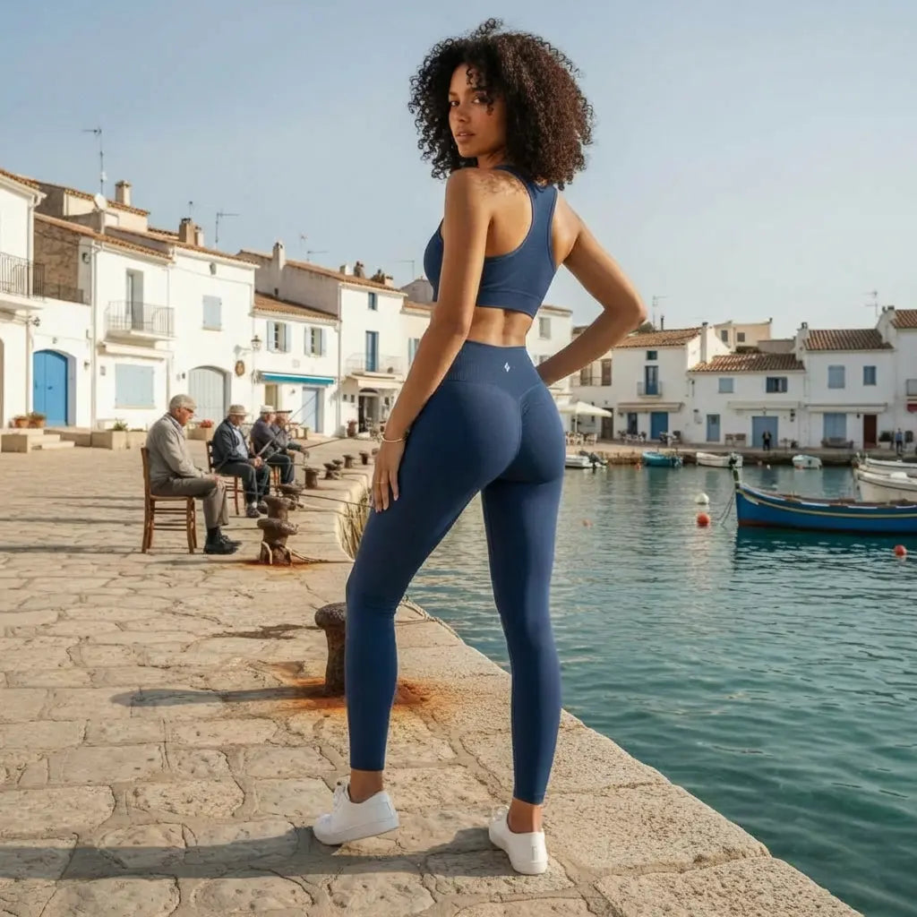 Une jeune femme aux cheveux bouclés pose dans une tenue de sport élégante composée du legging Elia bleu marine et d'un top coordonné. La scène se déroule par temps ensoleillé sur un quai en pierre bordant une eau turquoise, avec des maisons blanches traditionnelles et des bateaux de pêche en arrière-plan.