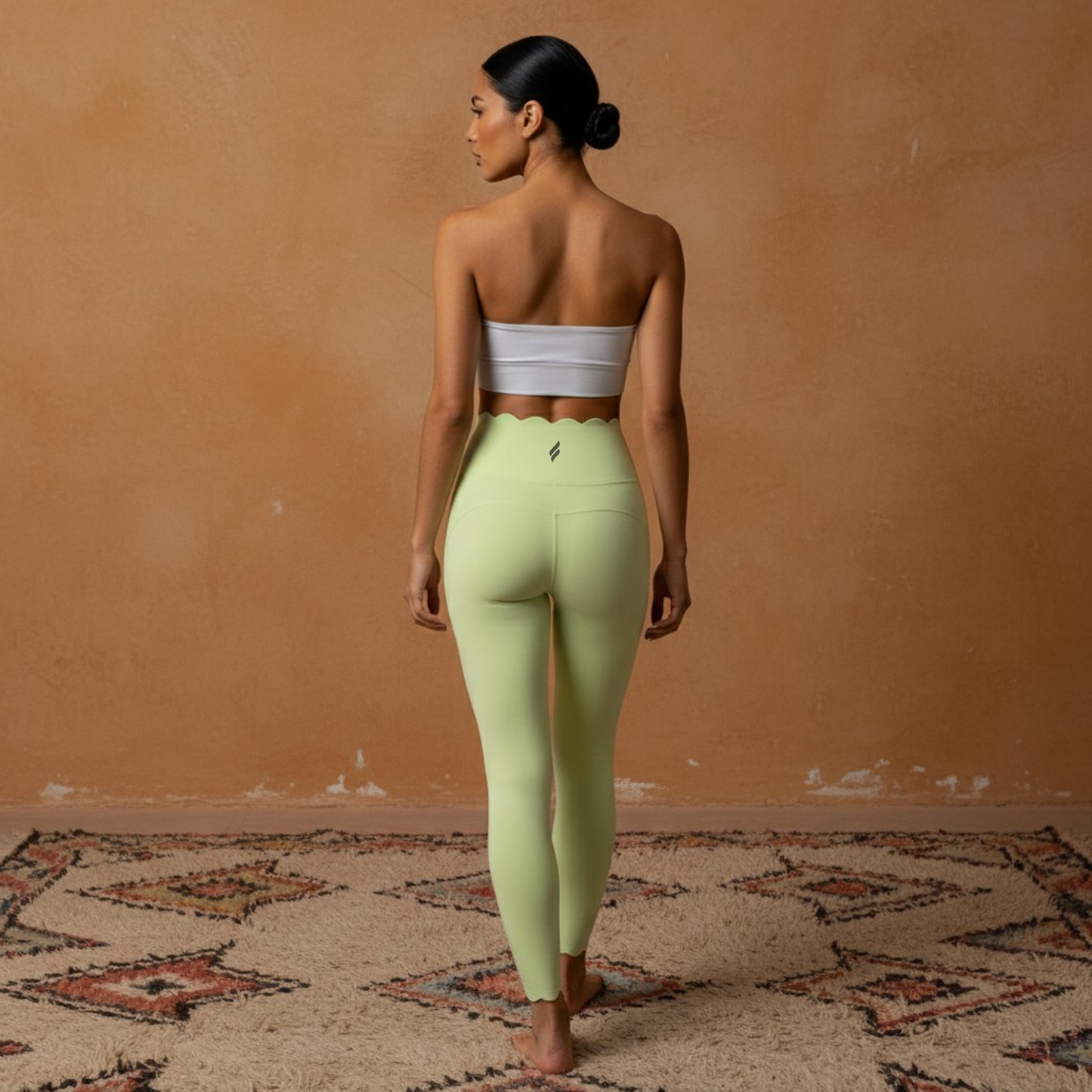 Une photographie de mode sportive montrant un legging de fitness taille haute de couleur vert menthe clair avec une finition festonnée distinctive à la taille. Le mannequin est de dos, portant un bandeau blanc minimaliste, debout sur un tapis à motifs géométriques devant un mur texturé ocre.