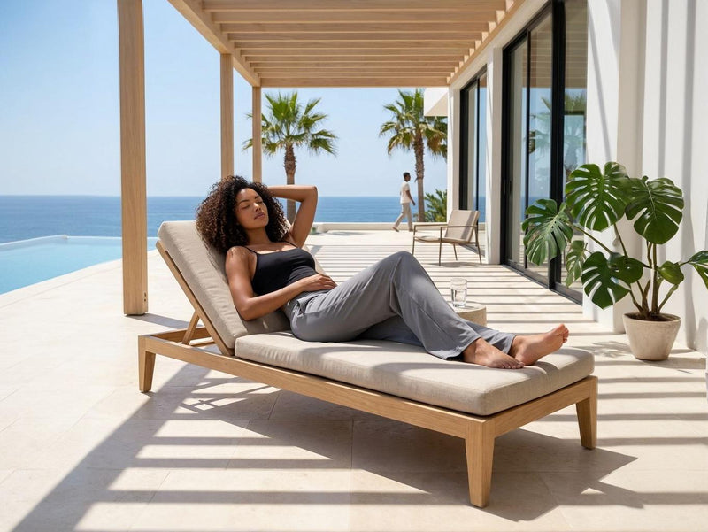 Une femme aux cheveux bouclés se détend sur une chaise longue moderne sous une pergola en bois. Elle porte un legging évasé gris de la marque Fyness, soulignant le confort et le style lifestyle. Le décor extérieur offre une vue panoramique sur l'océan avec une piscine à débordement et des palmiers.
