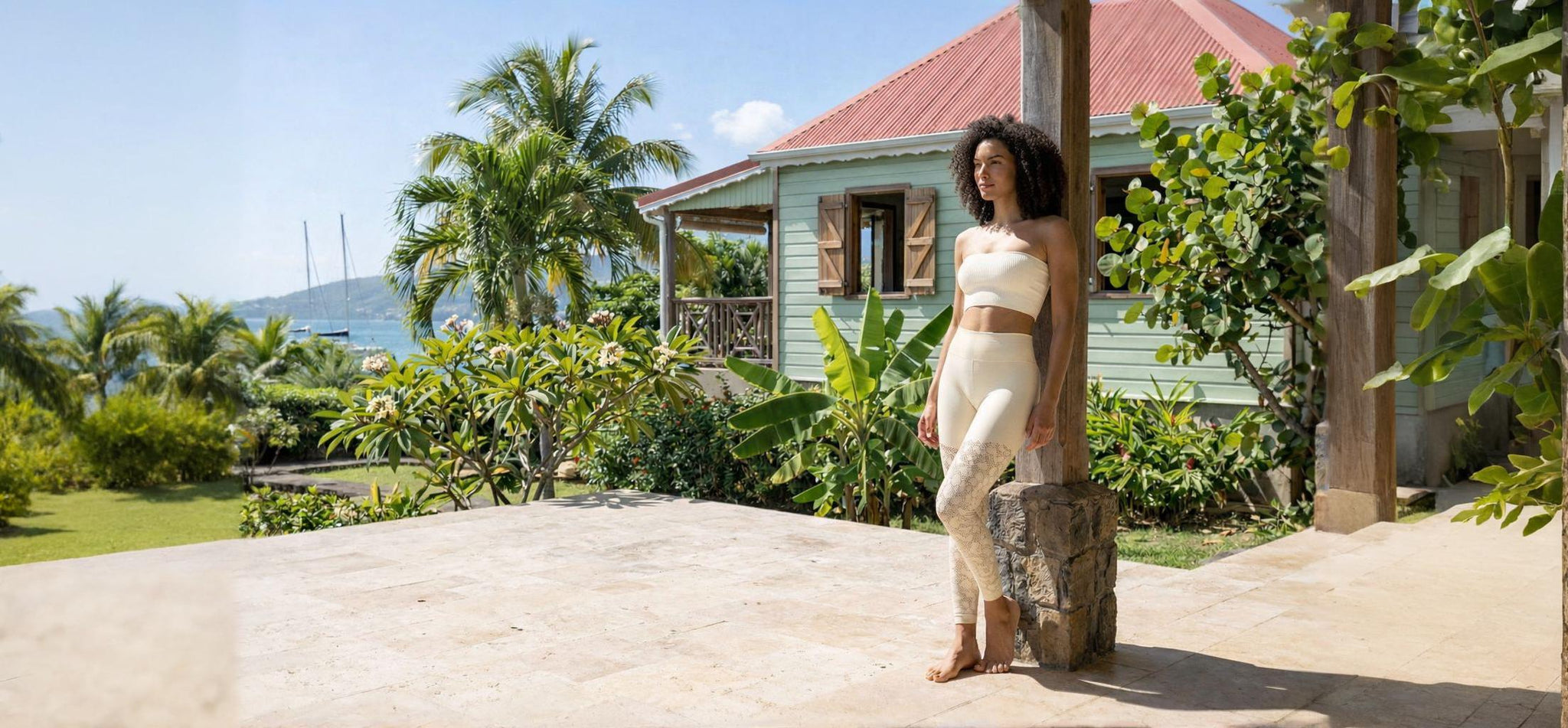 Une femme à la chevelure bouclée pose sur une terrasse en pierre devant une villa de style créole. Elle porte l'ensemble Kaia composé d'un legging galbant beige aux détails géométriques et d'un top bandeau côtelé, dans un environnement luxuriant avec des palmiers et des voiliers à l'horizon.