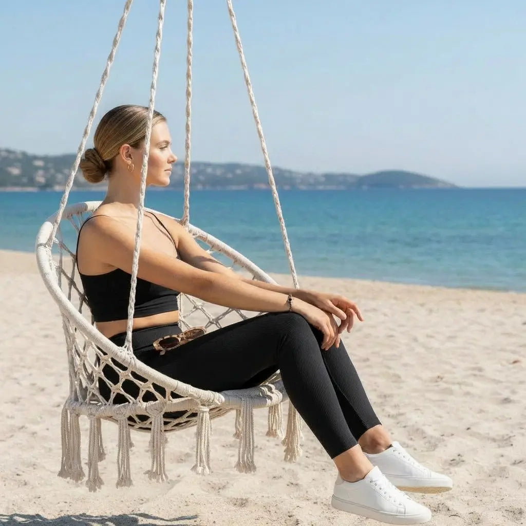 Une jeune femme pose de profil dans une chaise suspendue en macramé blanc sur une plage ensoleillée. Elle porte un legging noir ajusté et un haut court assorti, illustrant un style décontracté chic face à une mer bleue turquoise.
