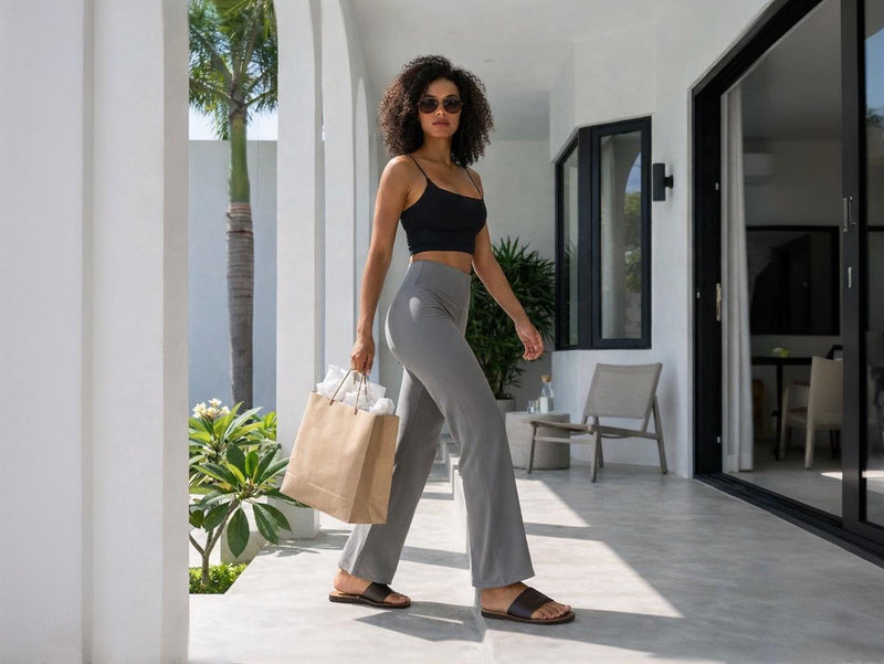 Une femme à la chevelure bouclée marche d'un pas assuré sur une terrasse ensoleillée aux arches blanches. Elle porte un legging de sport évasé gris ultra doux associé à un crop top noir et des lunettes de soleil, tenant un sac de shopping en papier brun pour un look lifestyle actif.