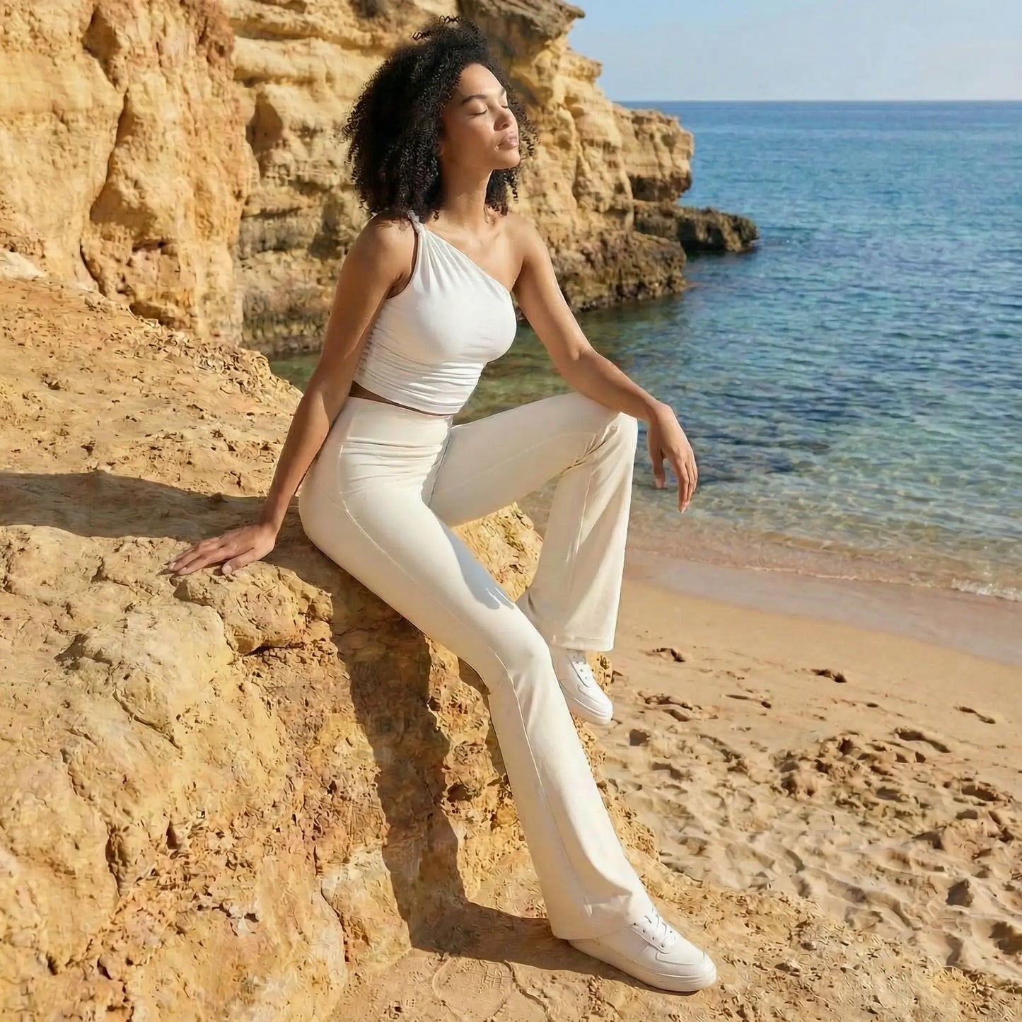 Une jeune femme à la chevelure bouclée pose sereinement sur un rocher doré surplombant une plage et une mer turquoise. Elle porte un ensemble de sport blanc immaculé comprenant un legging évasé taille haute et un haut à une seule épaule, créant un look athleisure élégant.
