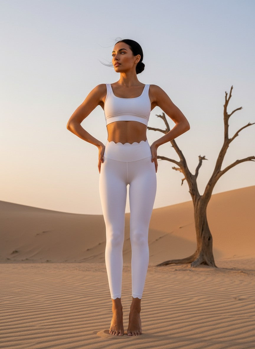 Une mannequin pose de face dans un décor de dunes de sable, vêtue d'une brassière et d'un legging de yoga blanc assorti. Le pantalon se distingue par ses finitions ondulées à la taille et aux chevilles, mis en valeur par la lumière dorée de fin de journée.