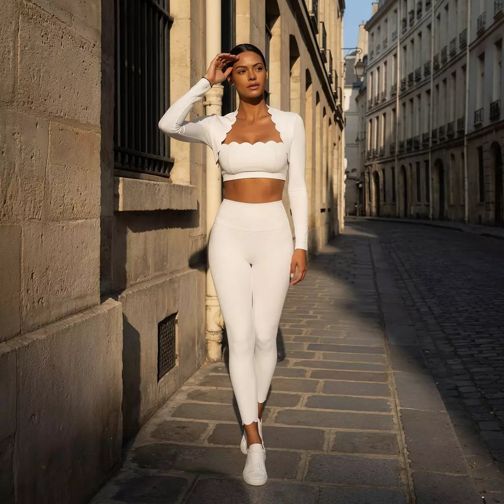 Une jeune femme marche avec élégance dans une rue pavée parisienne, vêtue d'un ensemble de sport blanc immaculé. La tenue se compose d'un legging haute performance, d'un crop top aux bords festonnés et d'un boléro assorti, le tout mis en valeur par une lumière dorée de fin de journée.