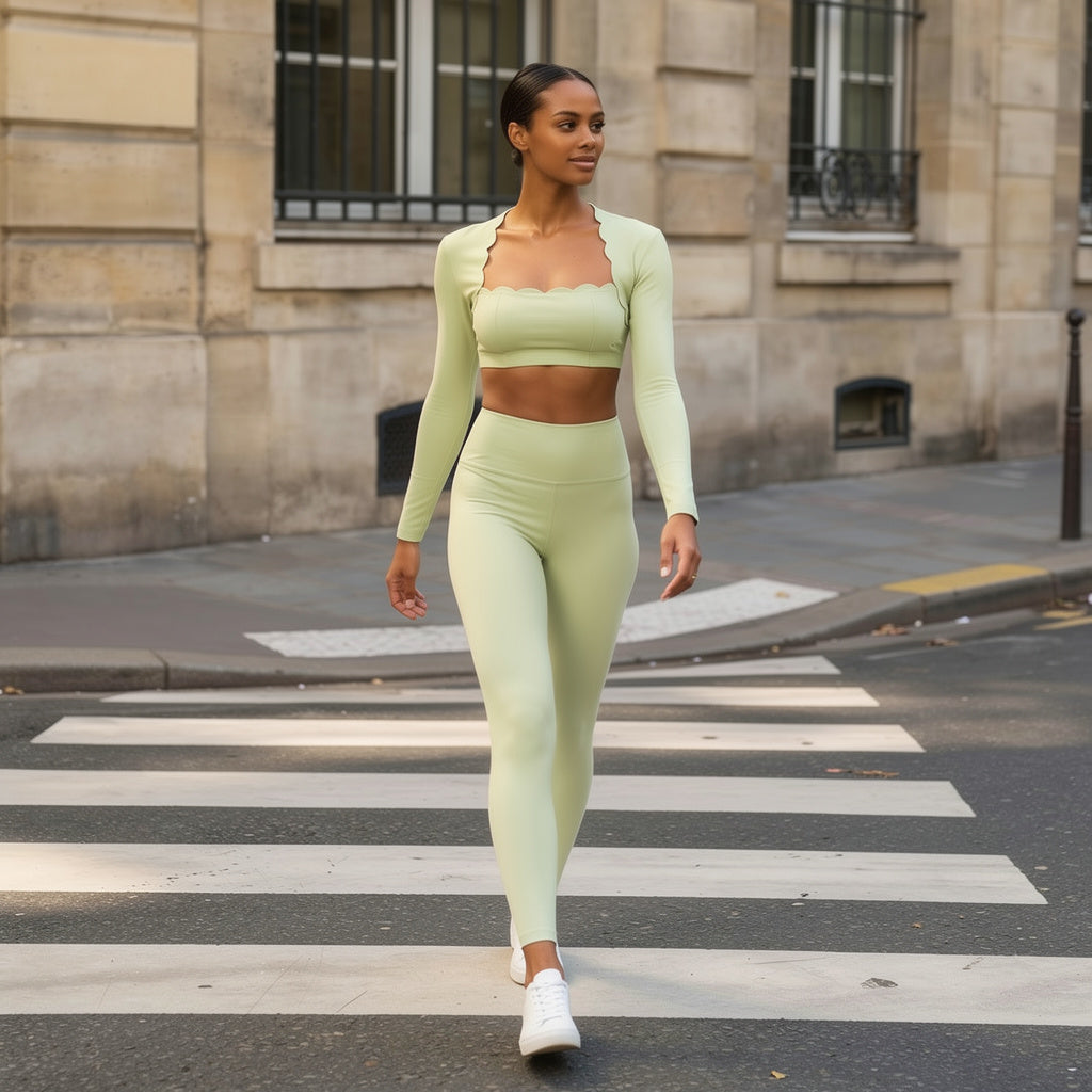 Une femme marche d'un pas assuré sur un passage piéton urbain, vêtue d'un ensemble de sport vert menthe sophistiqué. La tenue se compose d'un haut à manches longues aux finitions festonnées et d'un legging taille haute, mis en valeur par une architecture urbaine classique en arrière-plan.