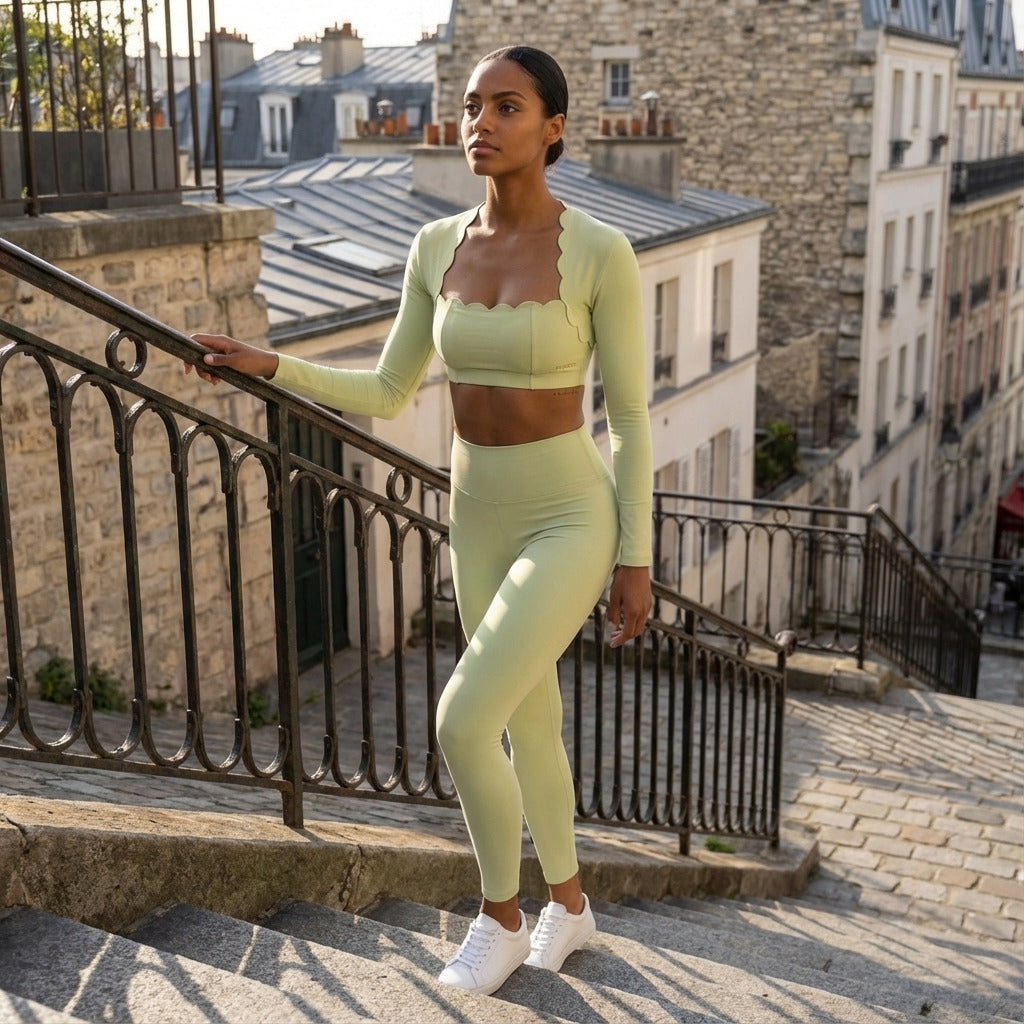 Une femme pose sur un escalier pavé parisien, vêtue d'un ensemble de sport vert sauge comprenant un legging taille haute et un top à manches longues aux finitions festonnées. La composition exploite les lignes diagonales de la rampe en fer forgé, mettant en valeur la silhouette dans une lumière naturelle douce sur fond d'architecture urbaine classique.