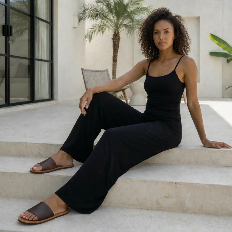 Une jeune femme pose avec élégance sur des escaliers extérieurs en pierre blanche, vêtue d'un ensemble monochrome noir composé d'un débardeur à fines bretelles et d'un legging évasé fluide. Elle complète son look minimaliste avec des sandales plates en cuir marron dans un décor épuré agrémenté d'un palmier.