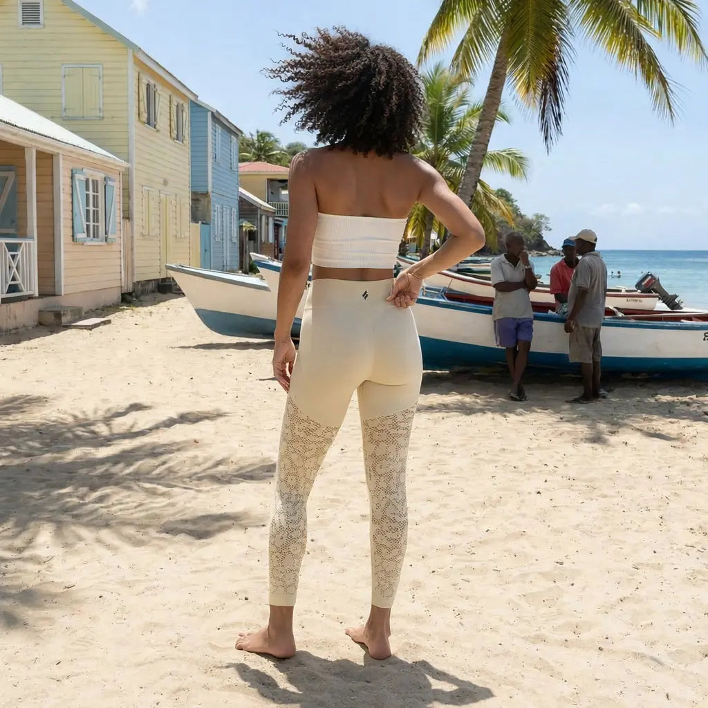 Une mise en situation lifestyle du legging galbant Kaia de la marque Fyness, mettant en avant sa coupe sculptante et ses motifs ajourés complexes sur le bas des jambes. La scène se déroule sur une plage authentique bordée de maisons colorées et de palmiers, soulignant l'esthétique élégante du vêtement.
