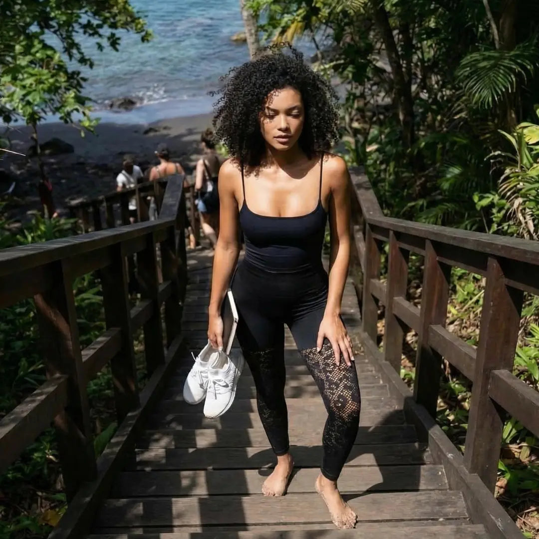 Une jeune femme aux cheveux bouclés porte une tenue de sport noire composée d'un débardeur et d'un legging galbant à motifs dentelés. Elle descend des marches en bois pieds nus, tenant ses baskets blanches et une tablette à la main, dans un cadre tropical bordé par l'océan.